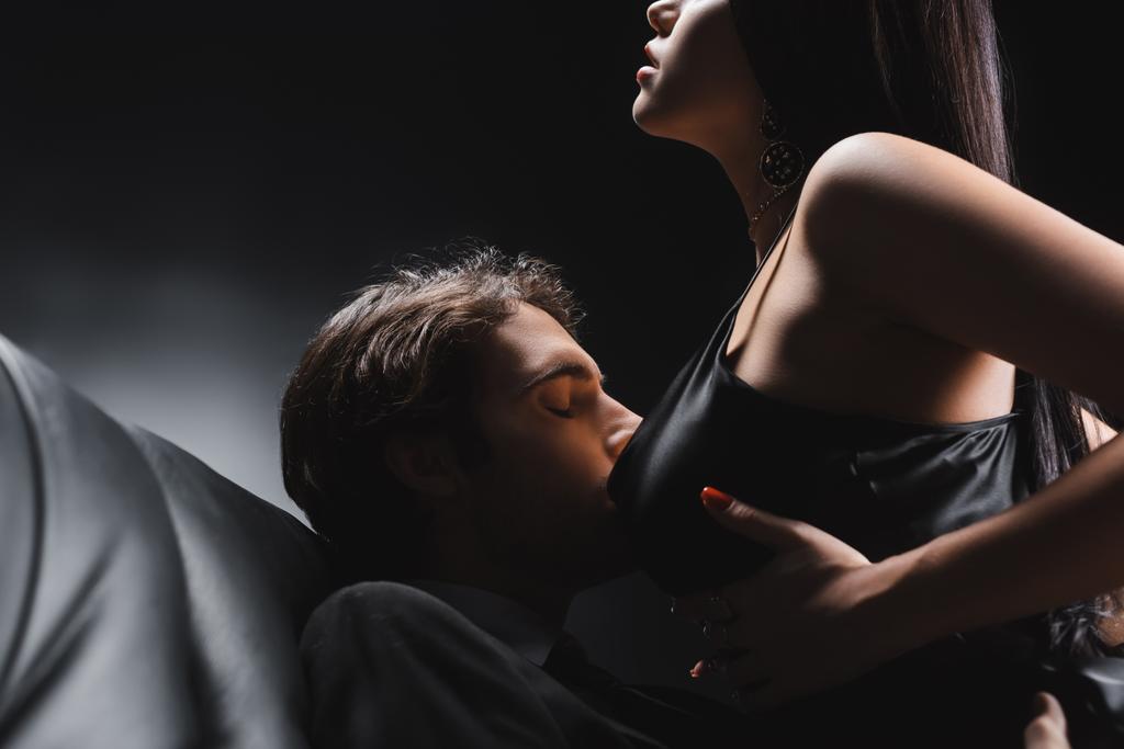 Young man kissing breast of passionate girlfriend on leather couch on black background - Fotó, kép