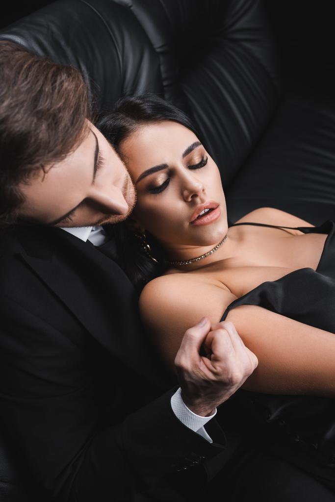 Hochwinkelaufnahme eines Mannes in formeller Kleidung, der Seidenkleid von einer sexy Freundin auf schwarzem Hintergrund auszieht - Foto, Bild