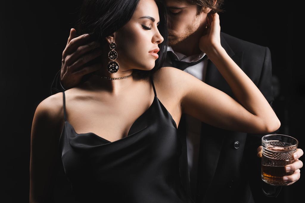 Öltönyös férfi kezében üveg whiskey és megható szexi nő selyem ruha elszigetelt fekete  - Fotó, kép