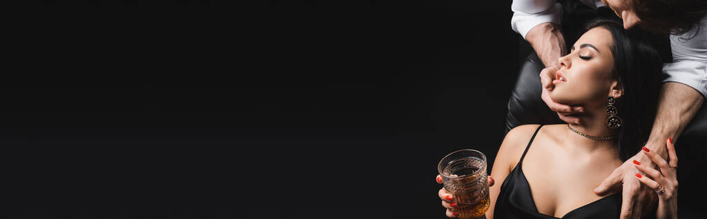 Νεαρός άνδρας αγγίζει αισθησιακή φίλη με ένα ποτήρι ουίσκι απομονωμένο σε μαύρο, banner  - Φωτογραφία, εικόνα