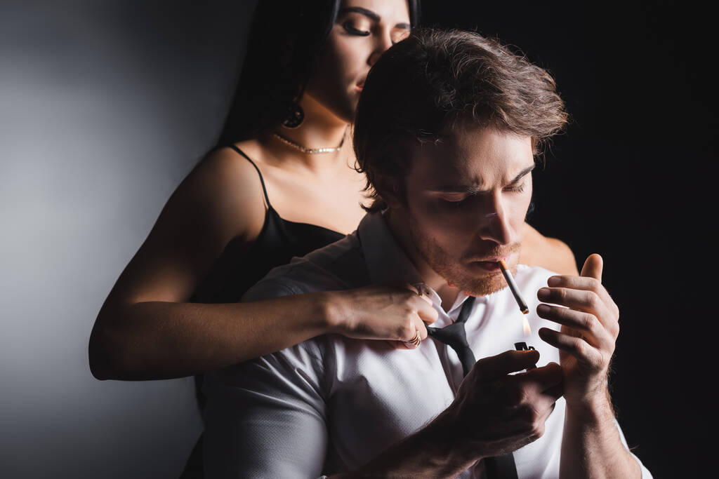 Passionate woman undressing boyfriend in shirt smoking cigarette on black background - Foto, Imagen