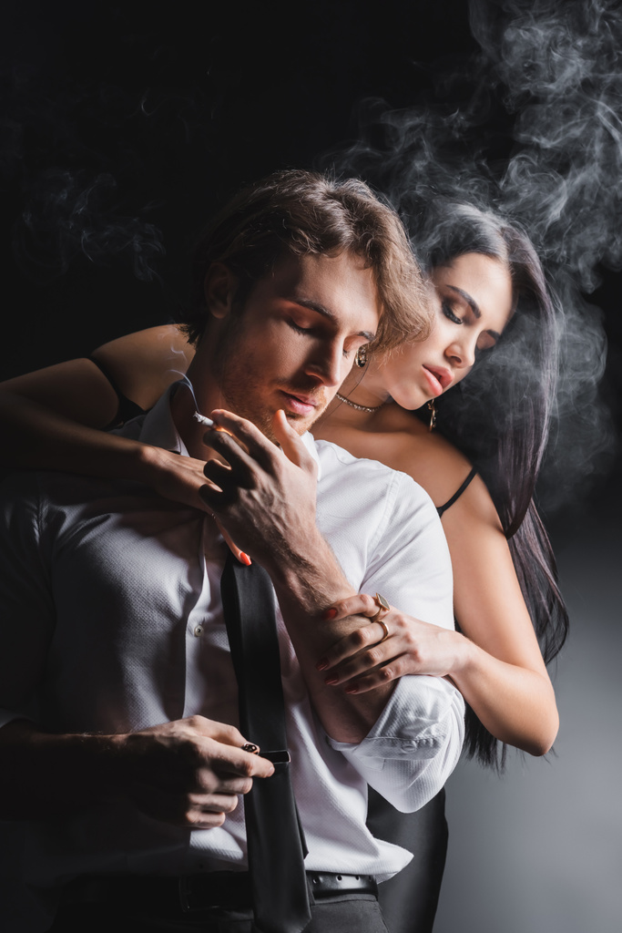 Sexy brunetka žena dotýkání přítel s cigaretou v blízkosti kouře na černém pozadí - Fotografie, Obrázek