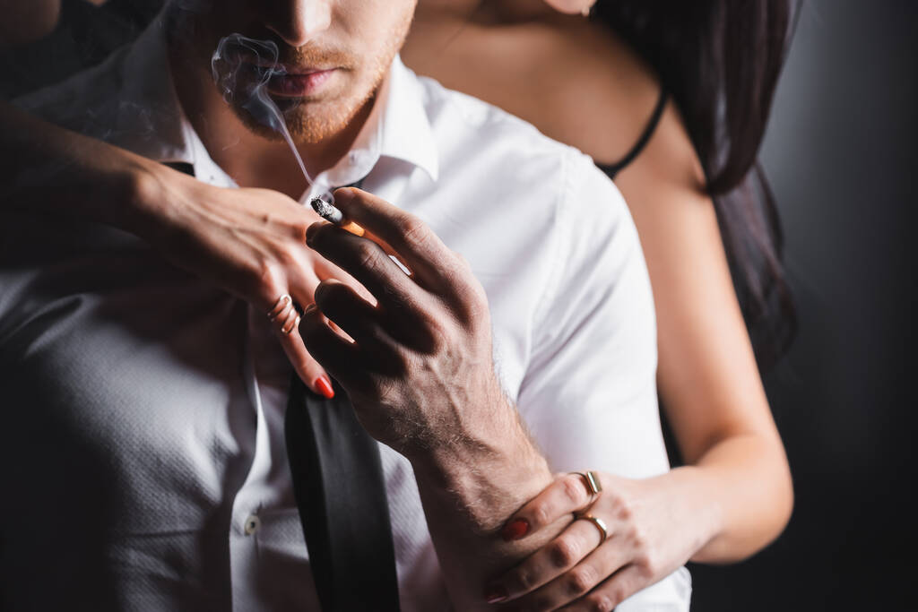 Ausgeschnittene Ansicht einer verschwommenen Frau, die Freund in formeller Kleidung mit Zigarette auf schwarzem Hintergrund berührt - Foto, Bild