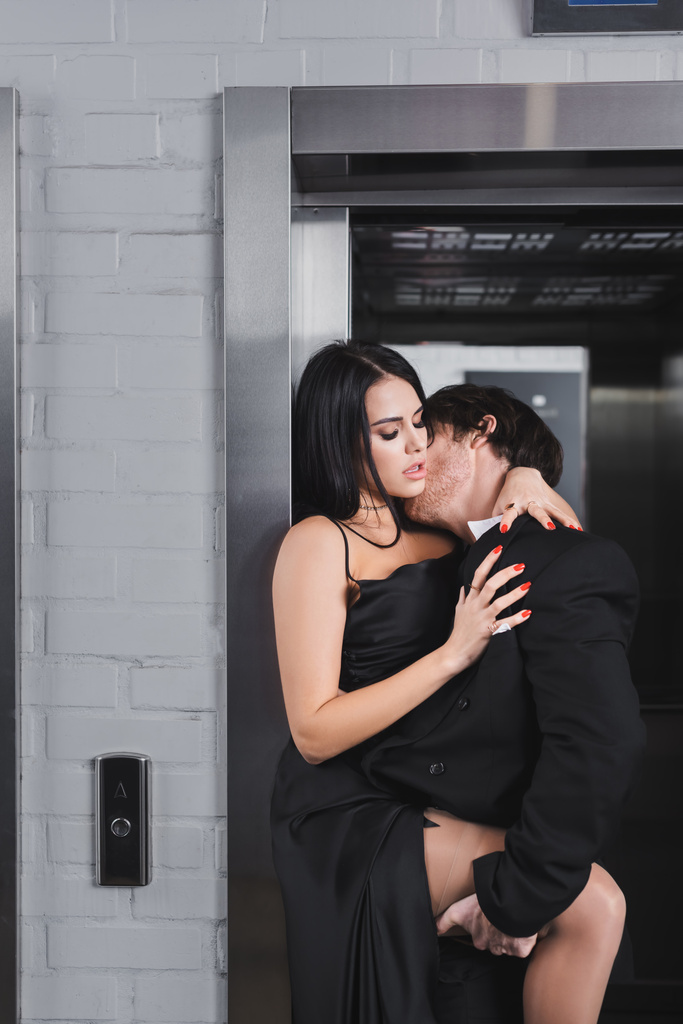 Чоловік Брюнет у костюмі цілує сексуальну жінку у сукні біля ліфта.  - Фото, зображення