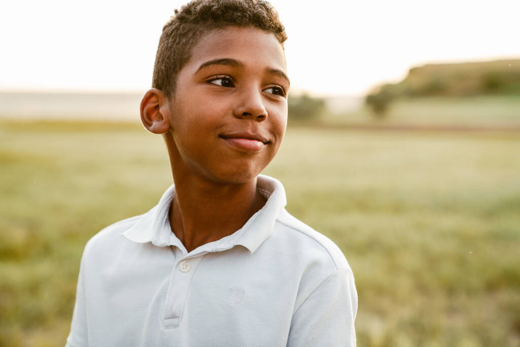 Beyaz tişörtlü siyah çocuk dışarıda poz verirken gülümsüyor ve yan bakıyor. - Fotoğraf, Görsel
