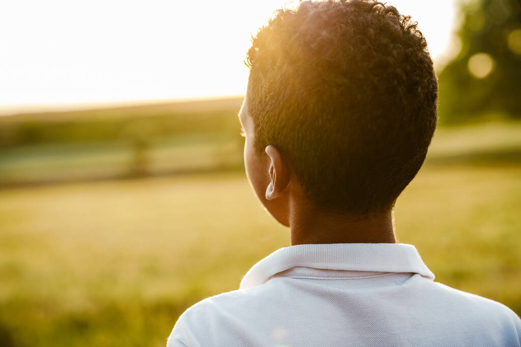 Μαύρο αγόρι φοράει λευκό πουκάμισο κοιτάζοντας το ηλιοβασίλεμα, ενώ στέκεται στο καλοκαιρινό πεδίο - Φωτογραφία, εικόνα