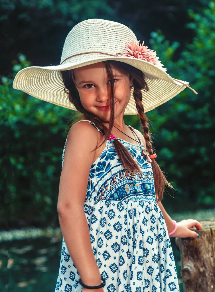 Beautiful little girl in a colorful dress with a hat on her head enjoys the summer day - Zdjęcie, obraz