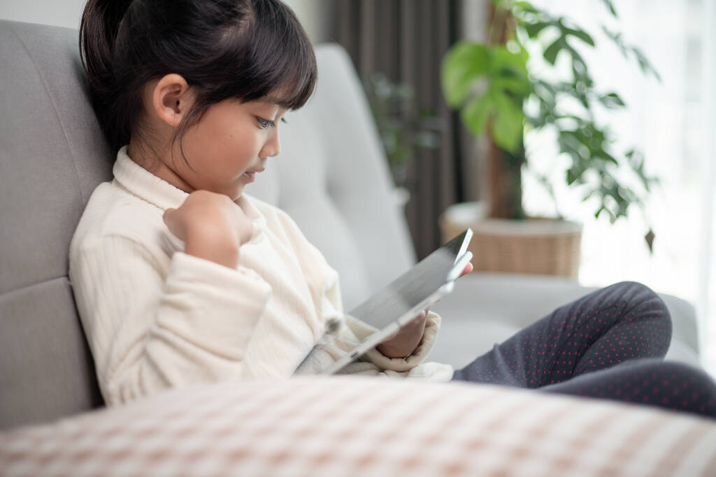 Dziewczynka korzystająca z tabletu w Internecie, dziecko siedzące na kanapie oglądające lub rozmawiające z przyjacielem online, dziecko relaksujące się rano w salonie, dzieci z koncepcją nowej technologii - Zdjęcie, obraz