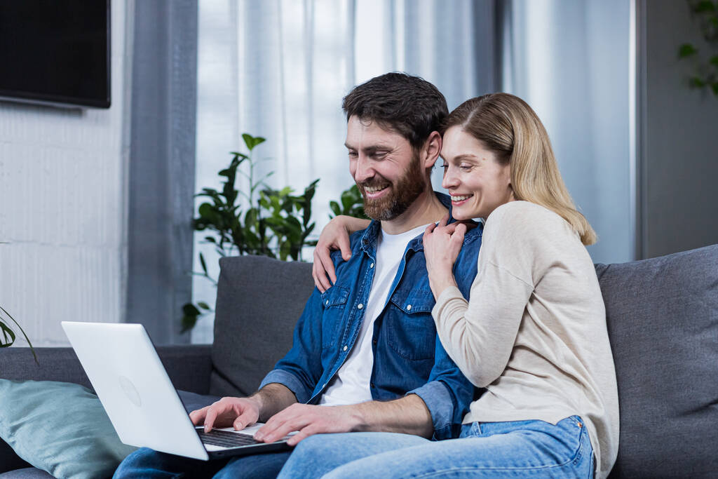 Szczęśliwe młode małżeństwo mężczyzna i kobieta razem w domu, siedzi na kanapie uśmiechnięty i za pomocą laptopa - Zdjęcie, obraz