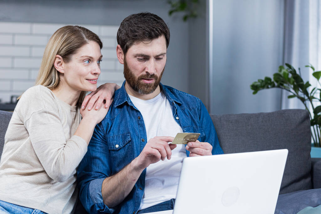 Happy married couple man and woman sitting at home on the couch using laptop for online shopping, holding a credit bank card - Photo, image