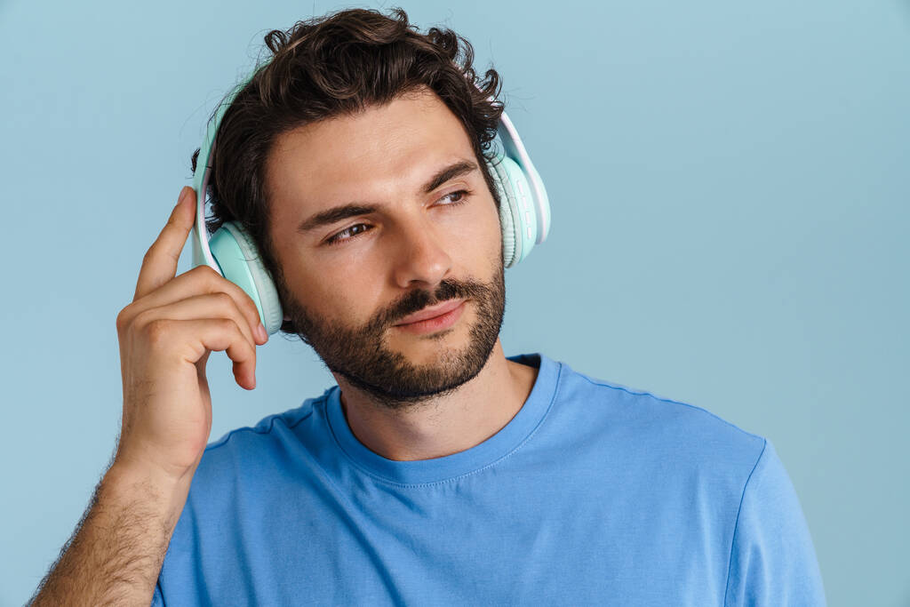Νεαρός μελαχρινός άντρας με μαλλιαρή ακουστική μουσική με ακουστικά απομονωμένα σε μπλε φόντο - Φωτογραφία, εικόνα