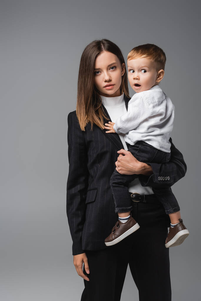 mujer en traje negro sosteniendo hijo sorprendido aislado en gris - Foto, imagen