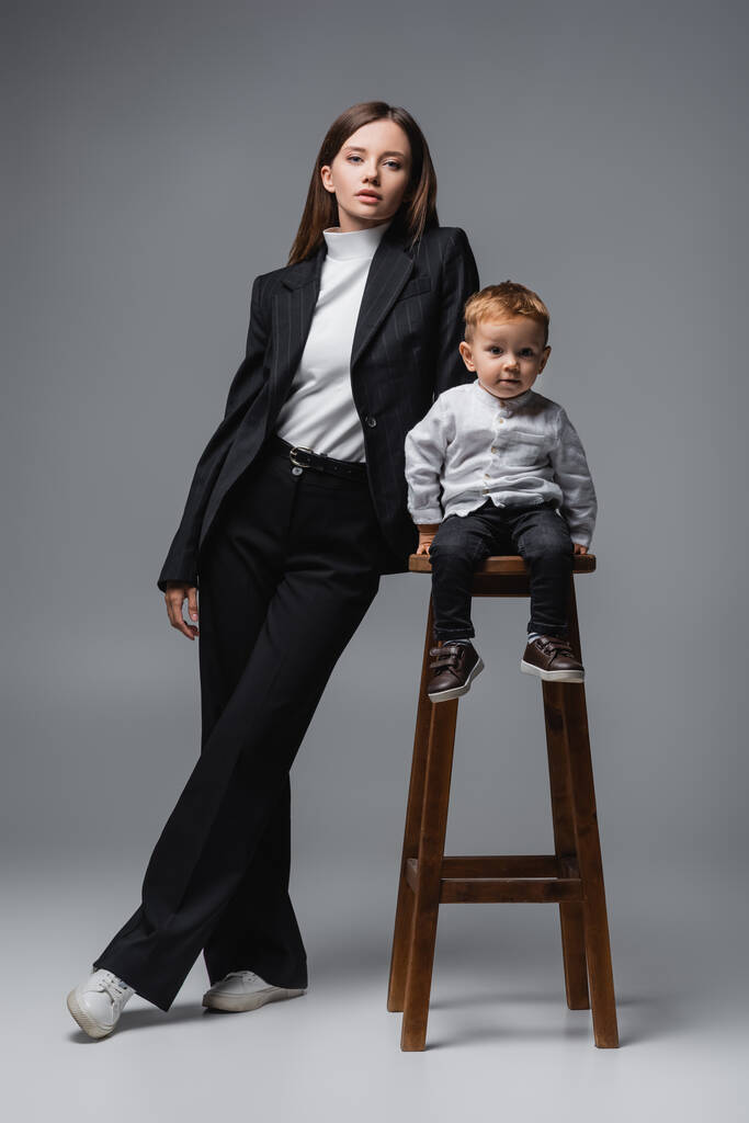 baby boy sitting on high stool and looking at camera near mom in black suit on grey - 写真・画像