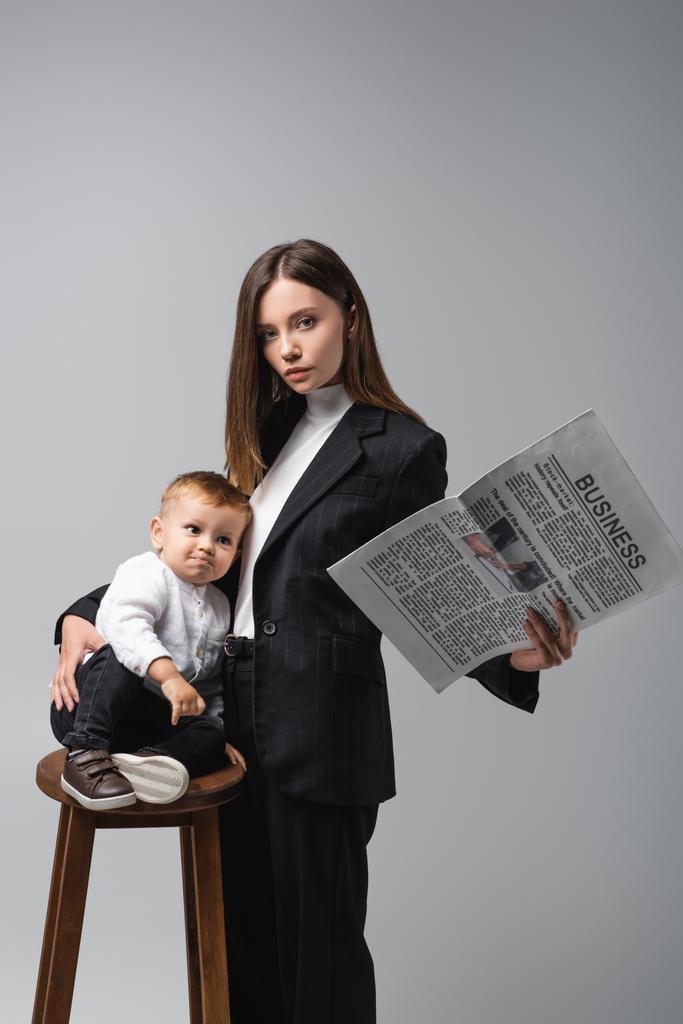 woman with business newspaper hugging son sitting on high stool isolated on grey - Photo, Image