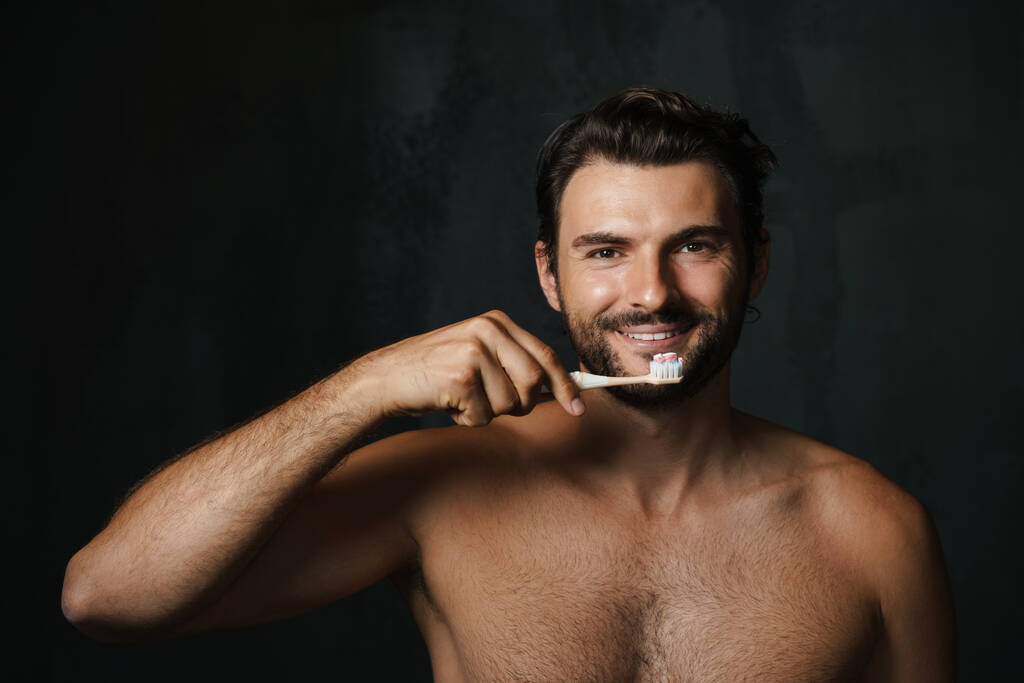 Νεαρός ημίγυμνος άντρας χαμογελάει ενώ βουρτσίζει τα δόντια του απομονωμένος σε μαύρο φόντο - Φωτογραφία, εικόνα