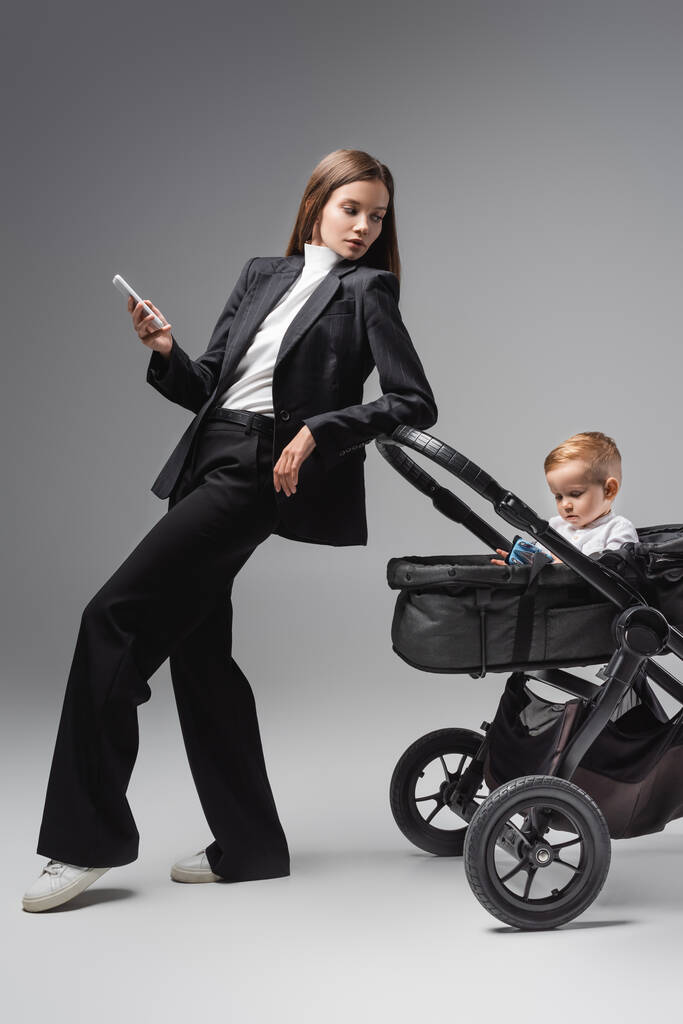 femme avec smartphone regardant son fils jouer avec une voiture jouet en landau sur gris - Photo, image