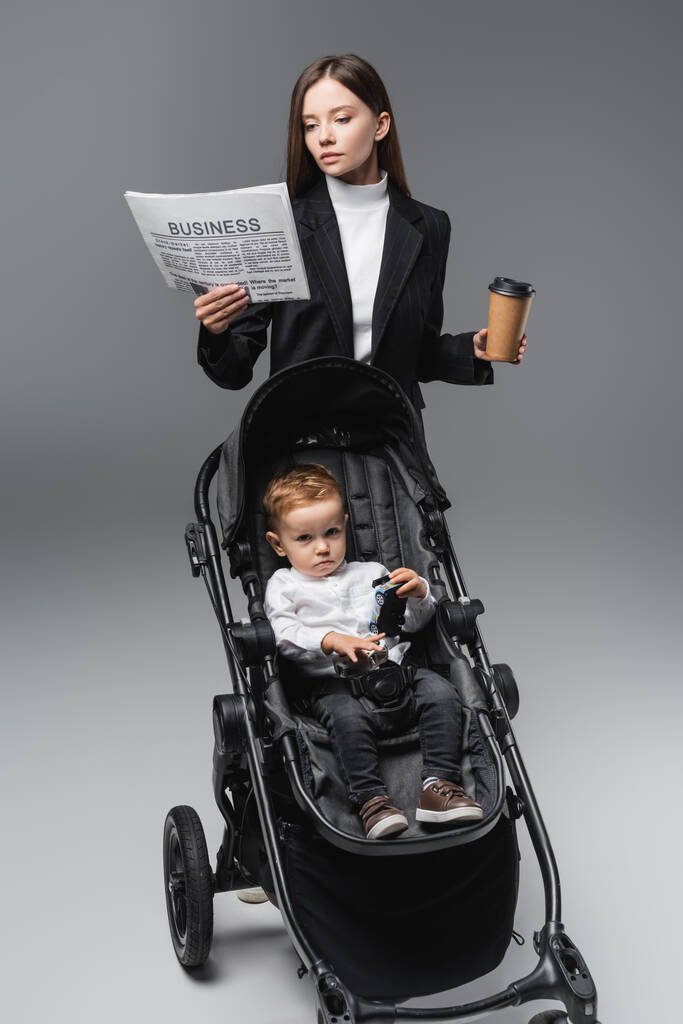 businesswoman with coffee to go reading newspaper near son in baby carriage on grey - Zdjęcie, obraz