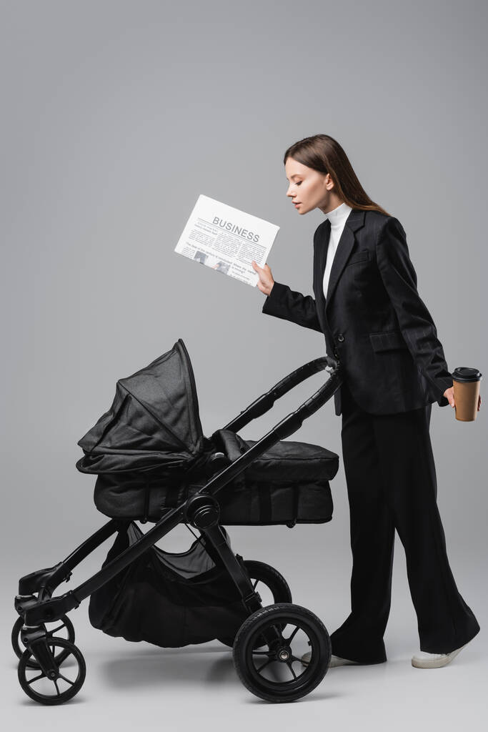 Seitenansicht einer Frau im Anzug mit Zeitung und Pappbecher in der Nähe eines schwarzen Kinderwagens auf grau - Foto, Bild