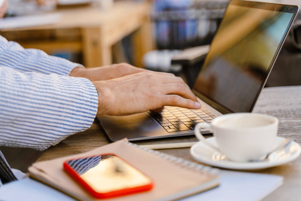 Młody mężczyzna w kurtce pracujący z laptopem podczas picia kawy w kawiarni na świeżym powietrzu - Zdjęcie, obraz
