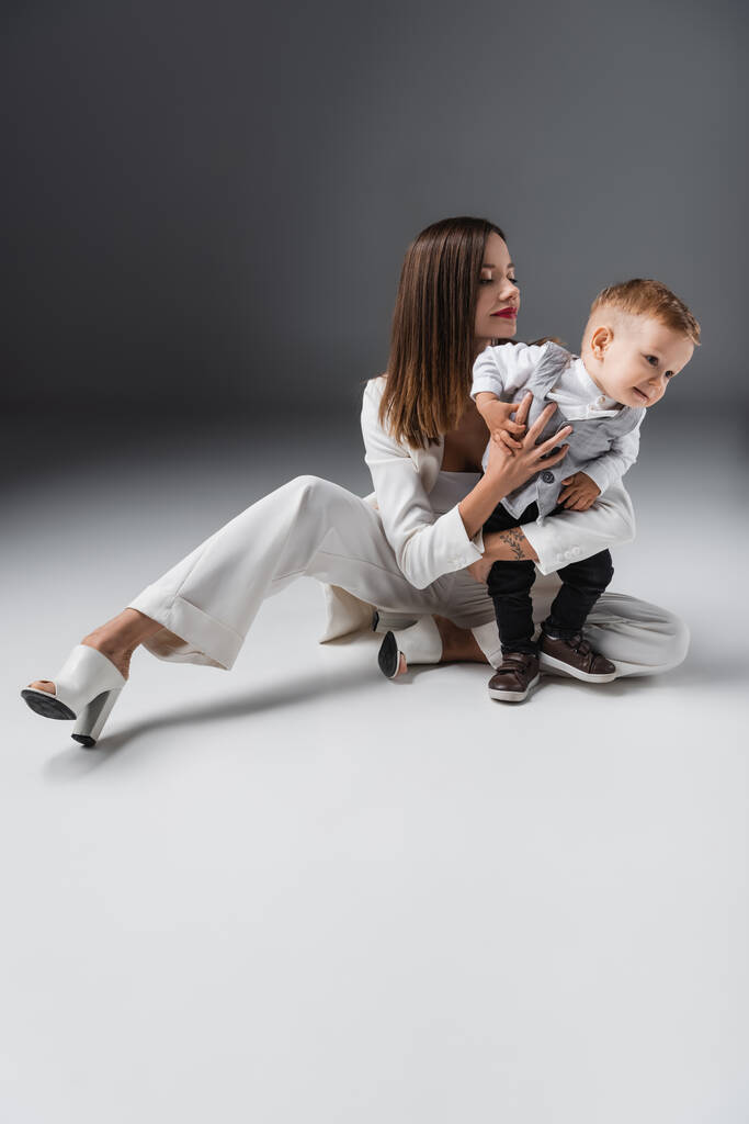 Ganzkörperansicht einer Frau im weißen Anzug, die ihren kleinen Sohn umarmt, während sie auf grau sitzt - Foto, Bild