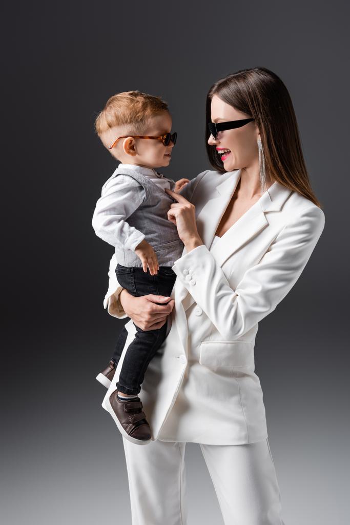 happy mother and son in trendy clothes and sunglasses looking at each other on grey - Foto, afbeelding