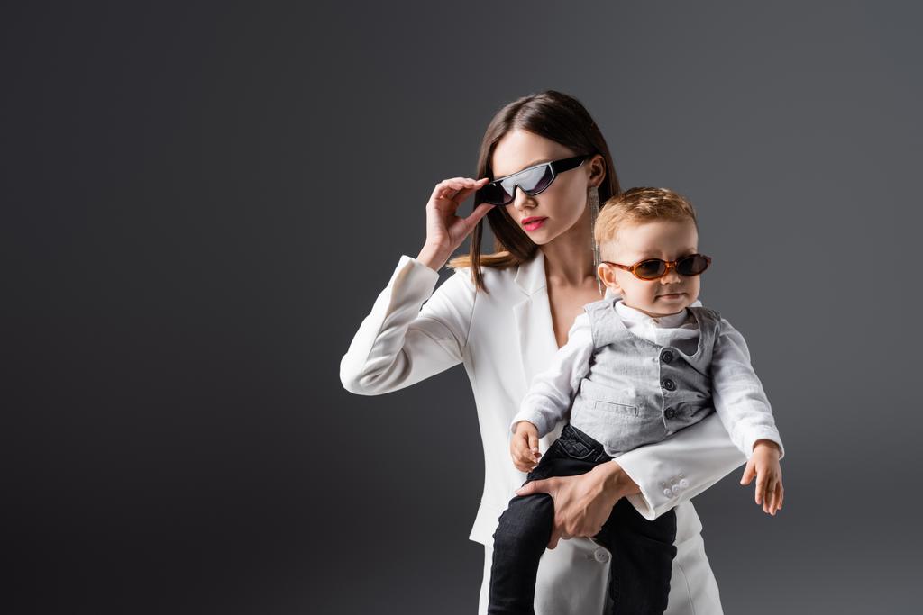 young woman adjusting stylish sunglasses while holding son isolated on grey - Fotoğraf, Görsel