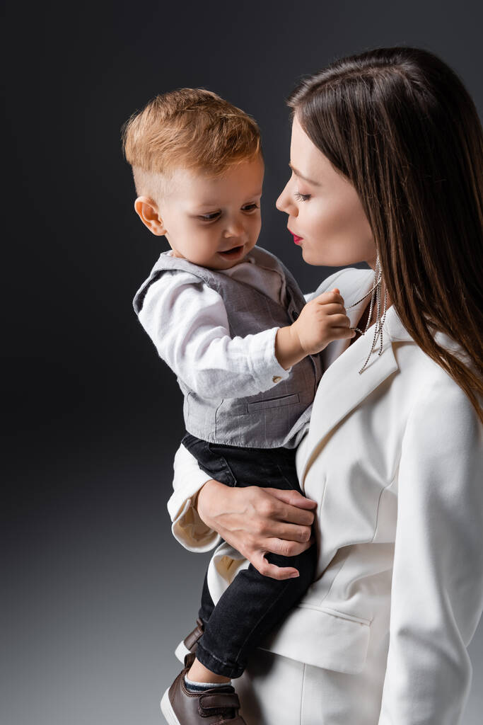 young and stylish woman holding son touching her necklace on grey - Foto, imagen