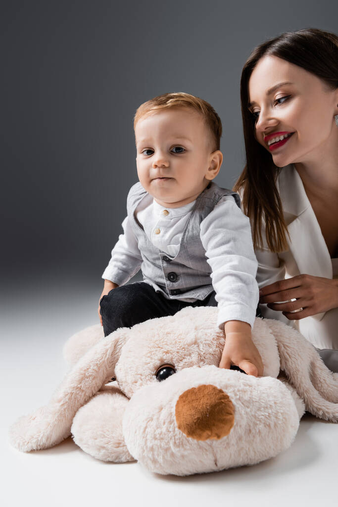 little boy looking at camera while sitting on huge toy dog near smiling mom on grey - Foto, Bild