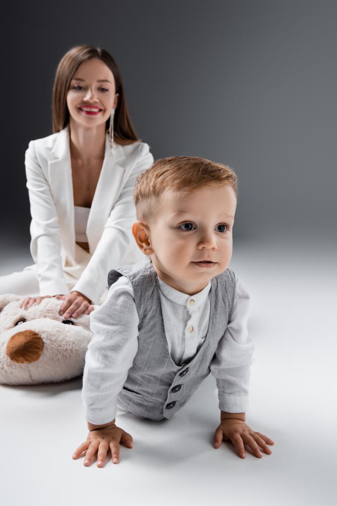 little boy in stylish clothes crawling near mother smiling on blurred background on grey - Photo, Image