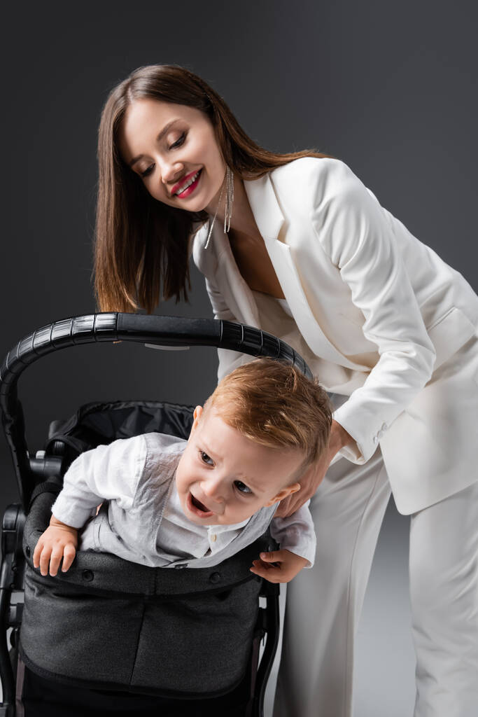 trendy woman smiling near excited son in baby carriage on grey - Foto, imagen