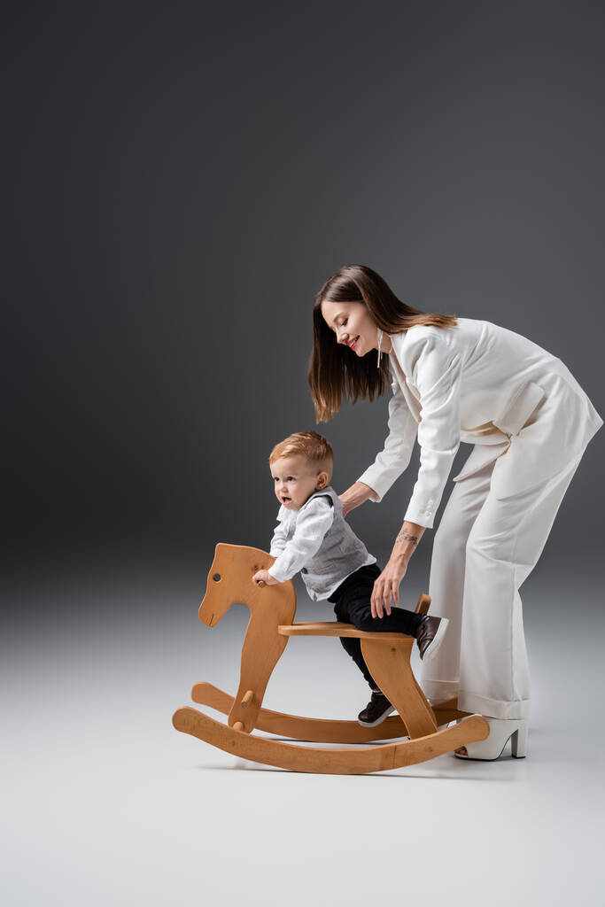 full length view of smiling woman supporting baby boy near rocking horse on grey - Photo, image