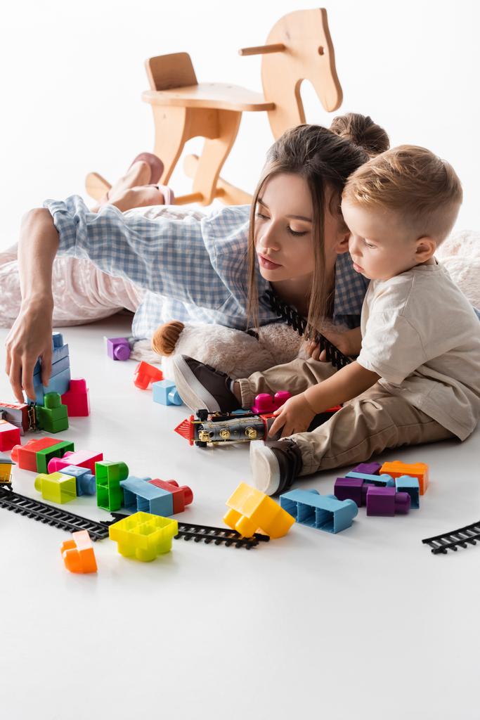 若い女性と幼児の男の子が白い上に建設キューブとおもちゃの鉄道で遊んでいます - 写真・画像
