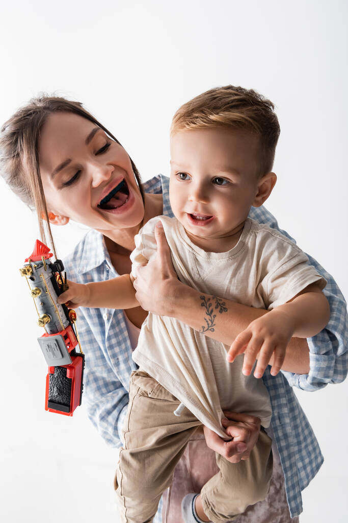 веселая женщина обнимает малыша сына держа игрушечный поезд изолирован на белом - Фото, изображение