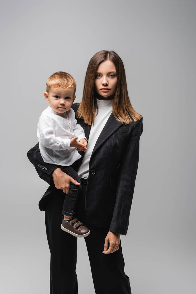 podnikatelka v obleku drží batole dítě a dívá se na kameru izolované na šedé - Fotografie, Obrázek