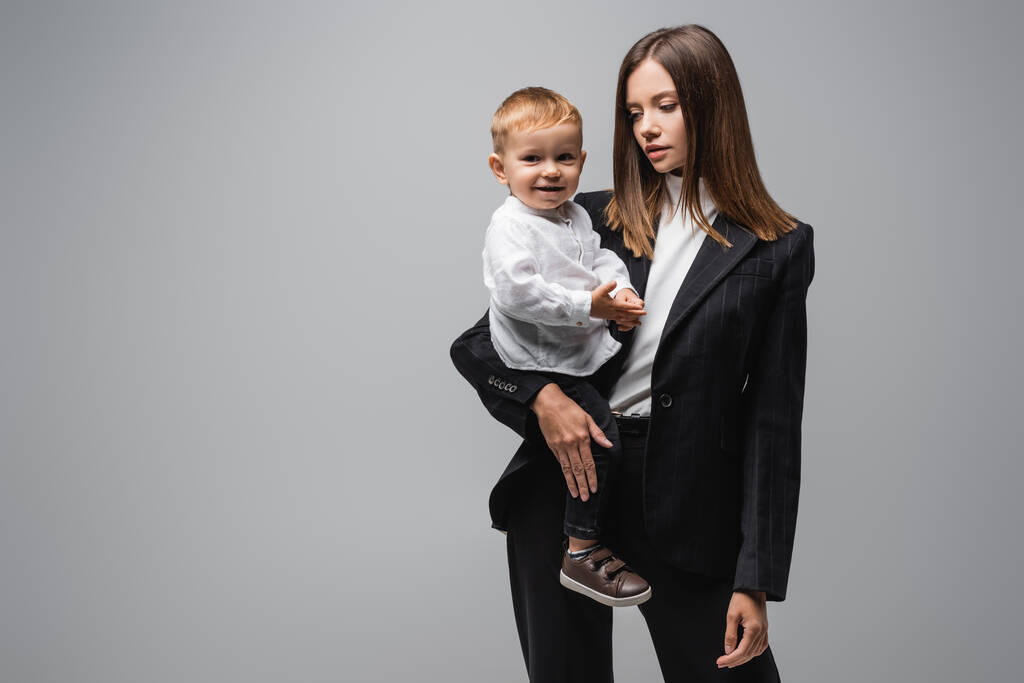 giovane donna d'affari che tiene piccolo figlio sorridente alla fotocamera isolata su grigio - Foto, immagini