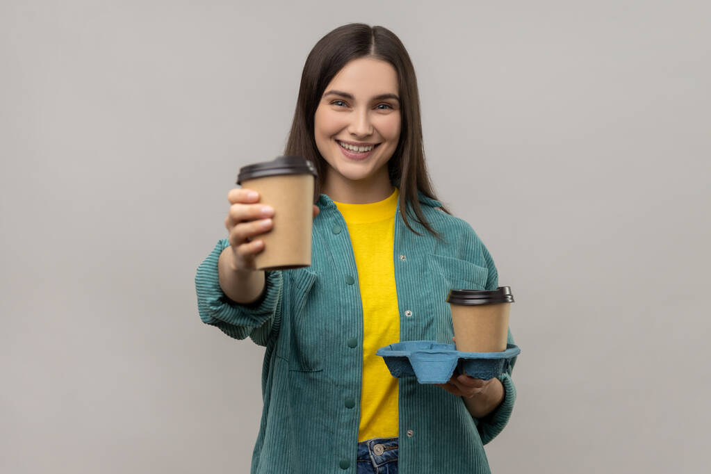 Portrét přátelské šťastné mladé ženy nabízí kávu, dává nápoje v jednorázové poháry a usmívá se, na sobě ležérní styl bundy. Vnitřní studio záběr izolované na šedém pozadí. - Fotografie, Obrázek