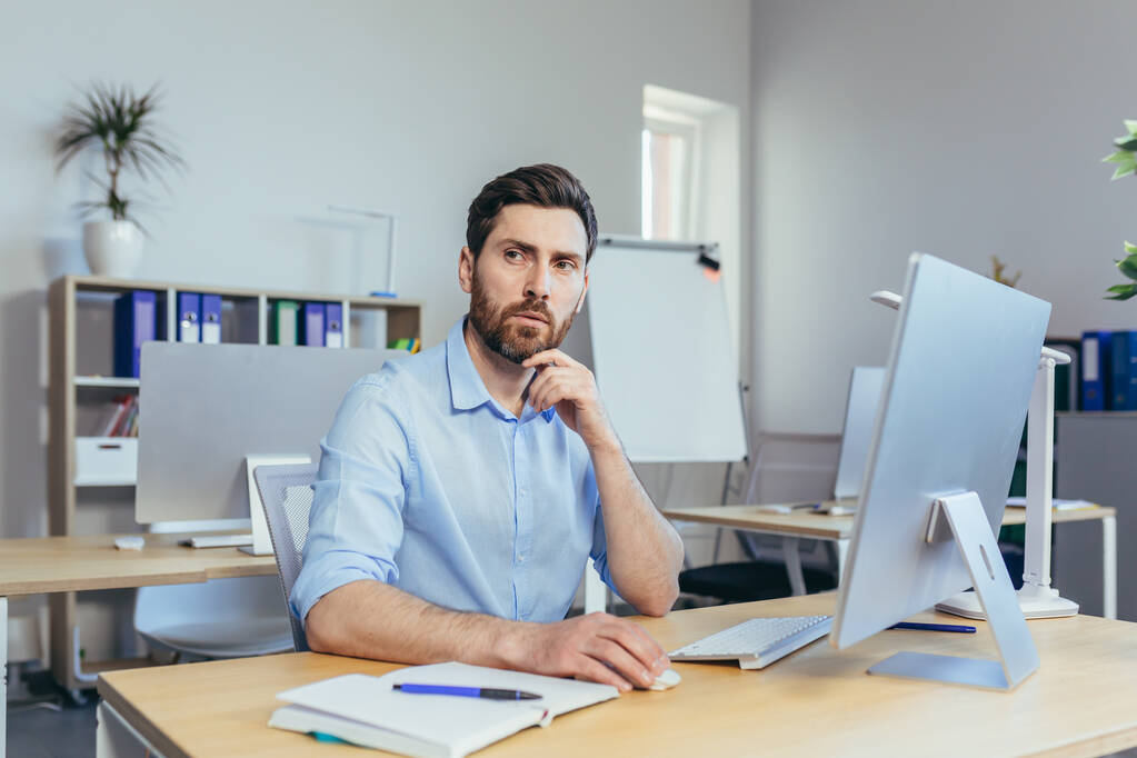 Portret van een serieuze en gefocuste zakenman, een man die overdag in een helder kantoor werkt, op de computer naar de monitor kijkend, in casual kleding - Foto, afbeelding