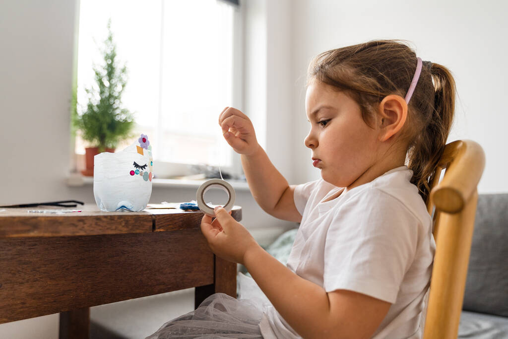 Bela menina pré-escolar caucasiana brincando em casa e fazendo artesanato de garrafa de plástico. Reciclar brinquedos e criatividade diy  - Foto, Imagem