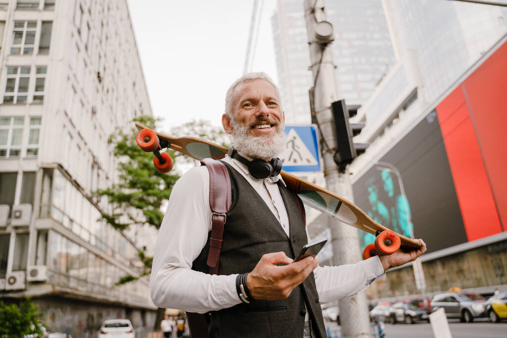 街の通りでスケートボードと一緒に立っている間、携帯電話を使用して灰色の成熟した男 - 写真・画像