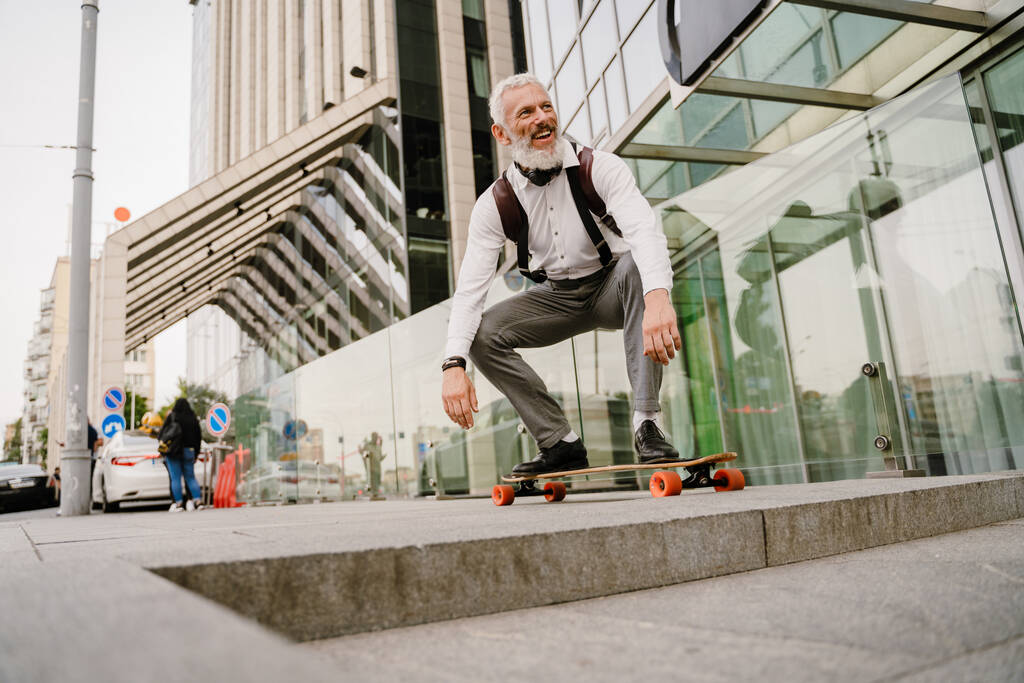 Сірий зрілий чоловік з бородою посміхається під час скейтбордингу на міській вулиці
 - Фото, зображення