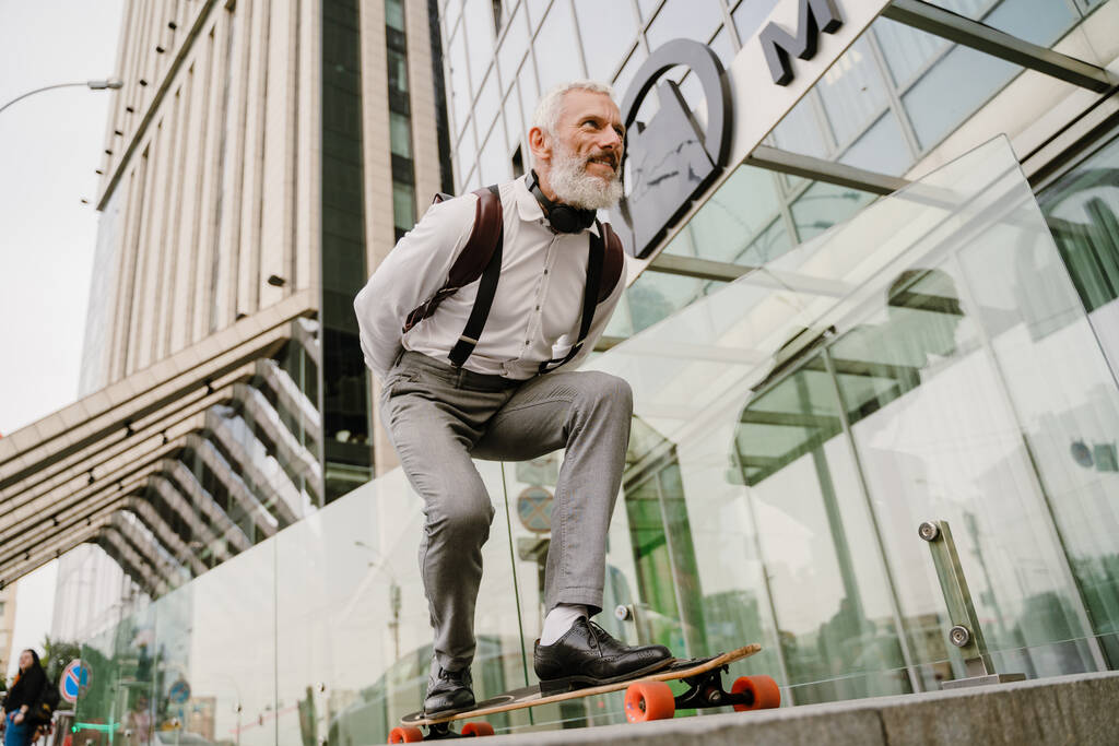 Серый взрослый мужчина с бородой, улыбающийся во время скейтбординга на городской улице - Фото, изображение
