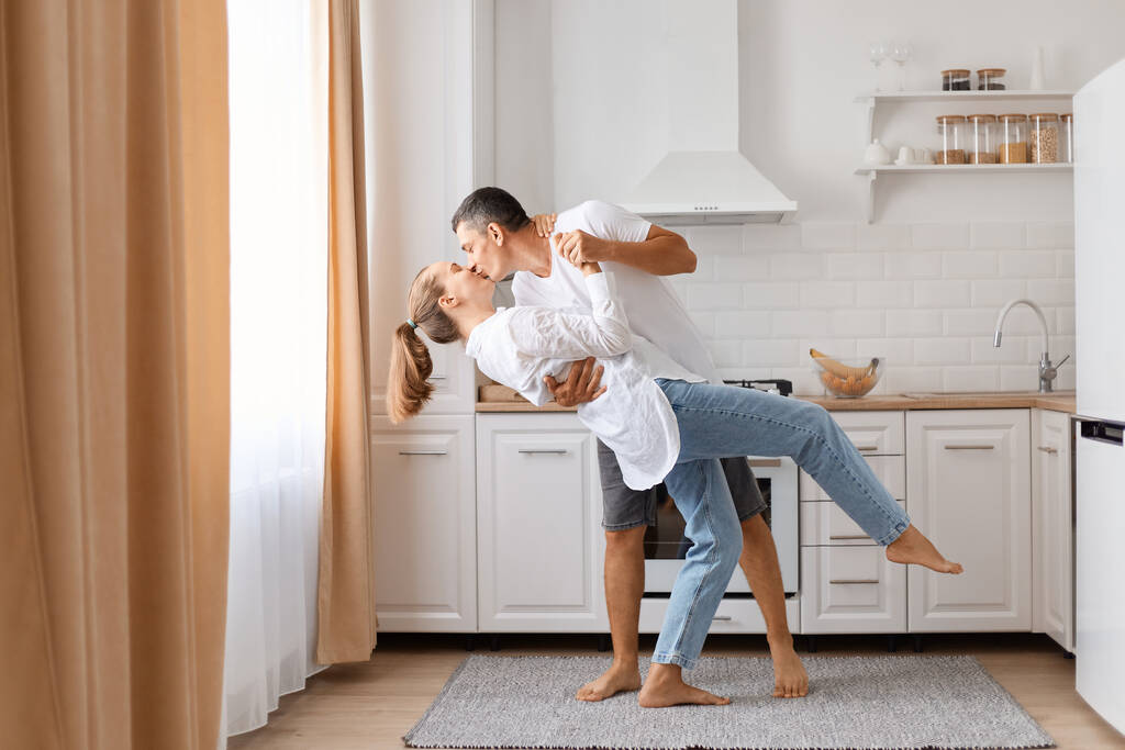 Indoor tiro de belo homem de cabelos escuros e atraente jovem mulher adulta se divertindo juntos na cozinha, casal romântico está dançando e beijando em casa. - Foto, Imagem