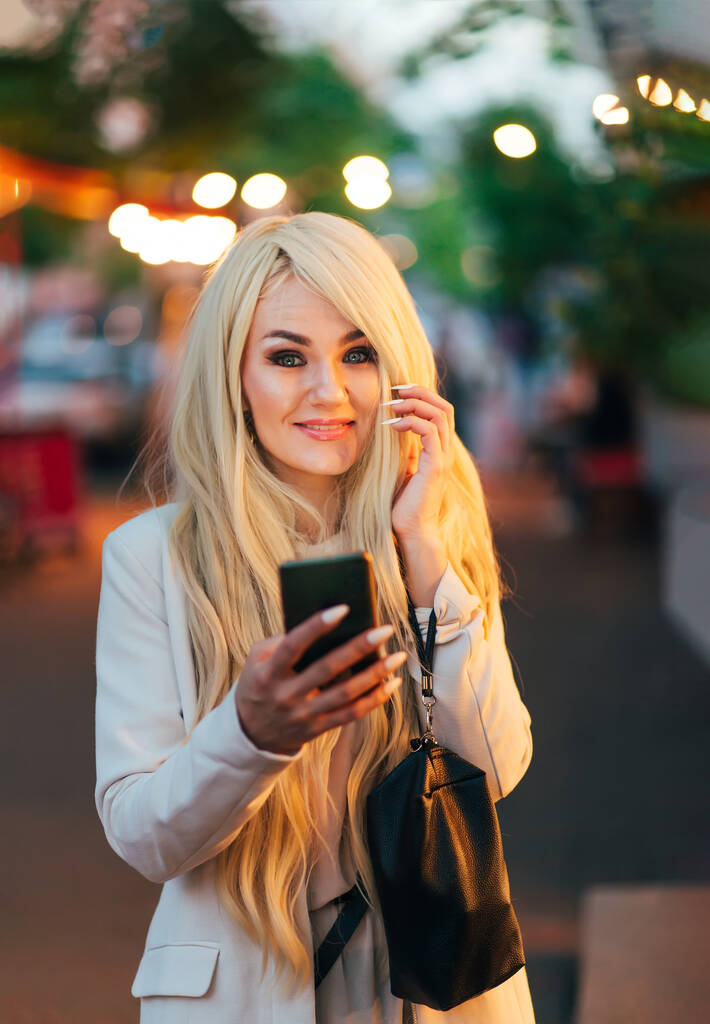 携帯電話を持ったライトジャケットを着て街の通りに金髪の美しい女性. - 写真・画像