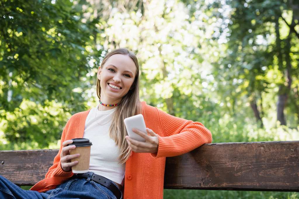 happy woman woman holding paper cup and smartphone while sitting on bench - Foto, imagen