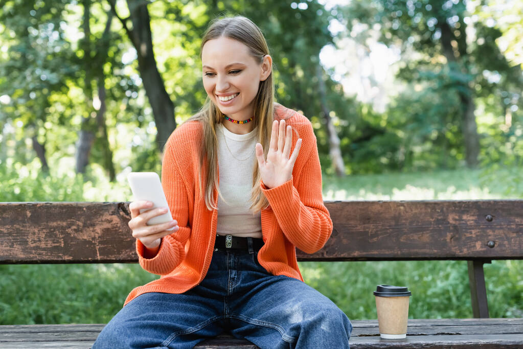весела жінка махає рукою під час відеодзвінка сидячи на лавці в зеленому парку
 - Фото, зображення