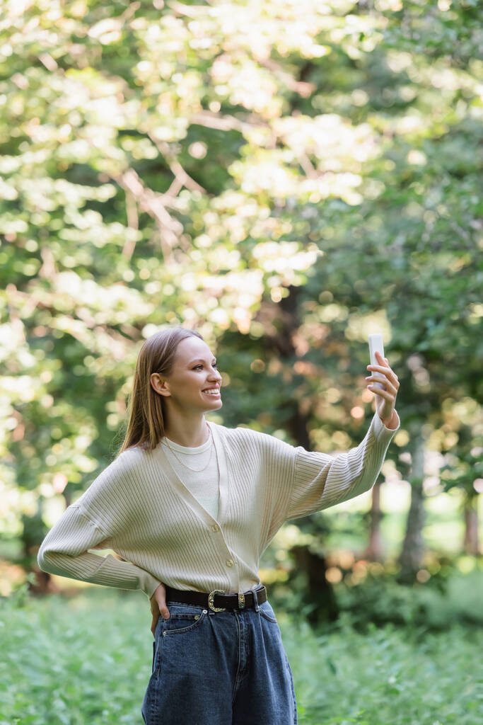 szczęśliwa młoda kobieta ze smartfonem stojącym z ręką na biodrze w parku  - Zdjęcie, obraz
