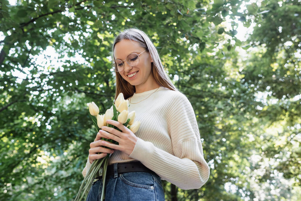 low angle view of happy woman in glasses holding tulips in green park - Fotó, kép
