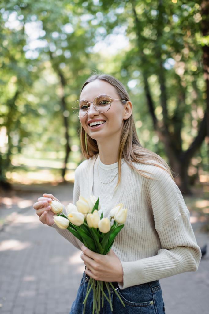 cheerful woman in glasses holding bouquet of tulips in green park - Fotoğraf, Görsel