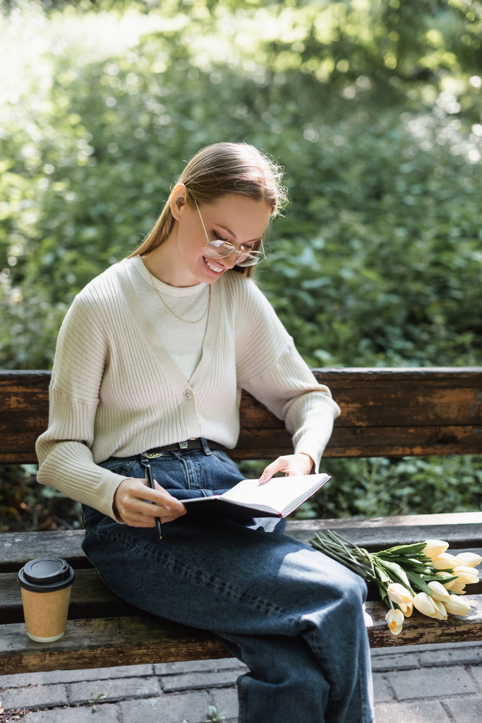 happy woman in jeans holding notebook near bouquet of tulips and paper cup on bench - Foto, Imagen