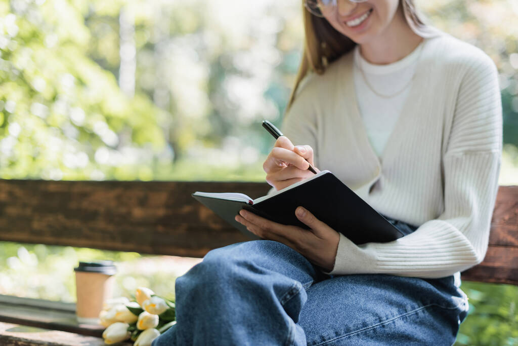 przycięty widok wesołej kobiety piszącej w notatniku i siedzącej na ławce - Zdjęcie, obraz